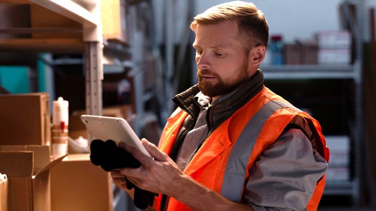 Arbeiter in Warnweste mit Tablet in der Hand vor Lagerregalen