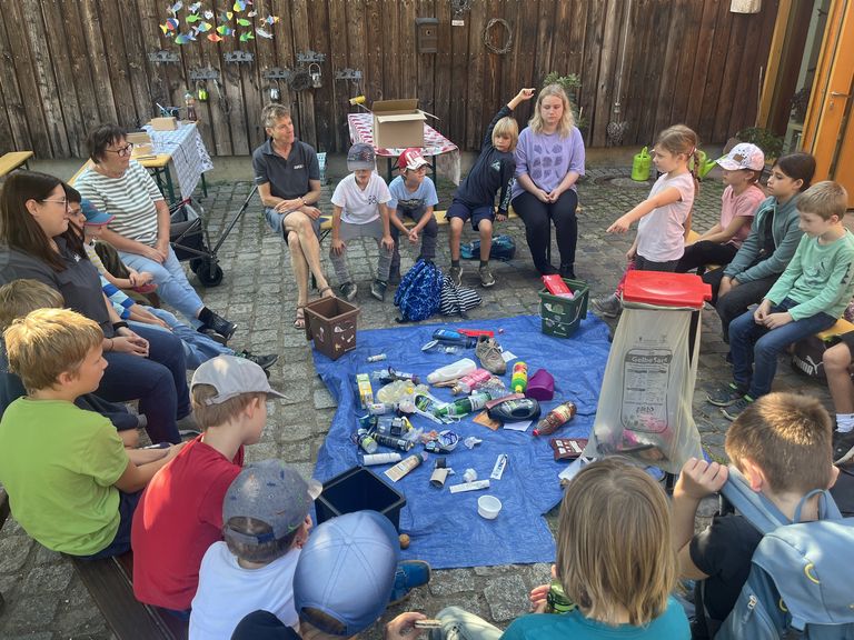 Kinder und Erwachsene sitzen im Kreis um eine Decke mit Abfällen