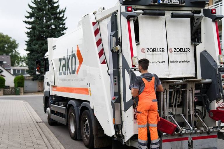 Lader in oranger Sicherheitskleidung steht hinten auf dem Trittbrett eines Abfallsammelfahrzeugs